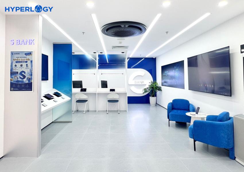 261019 sự kiện Smart banking 03 - Hyperlogy Modern bank lobby with white desks, blue accents, a circular BANK logo, and blue armchairs against wall screens in a bright space.